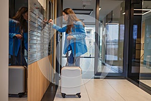 Woman with a suitcase opening mailbox in hallway of apartment building 